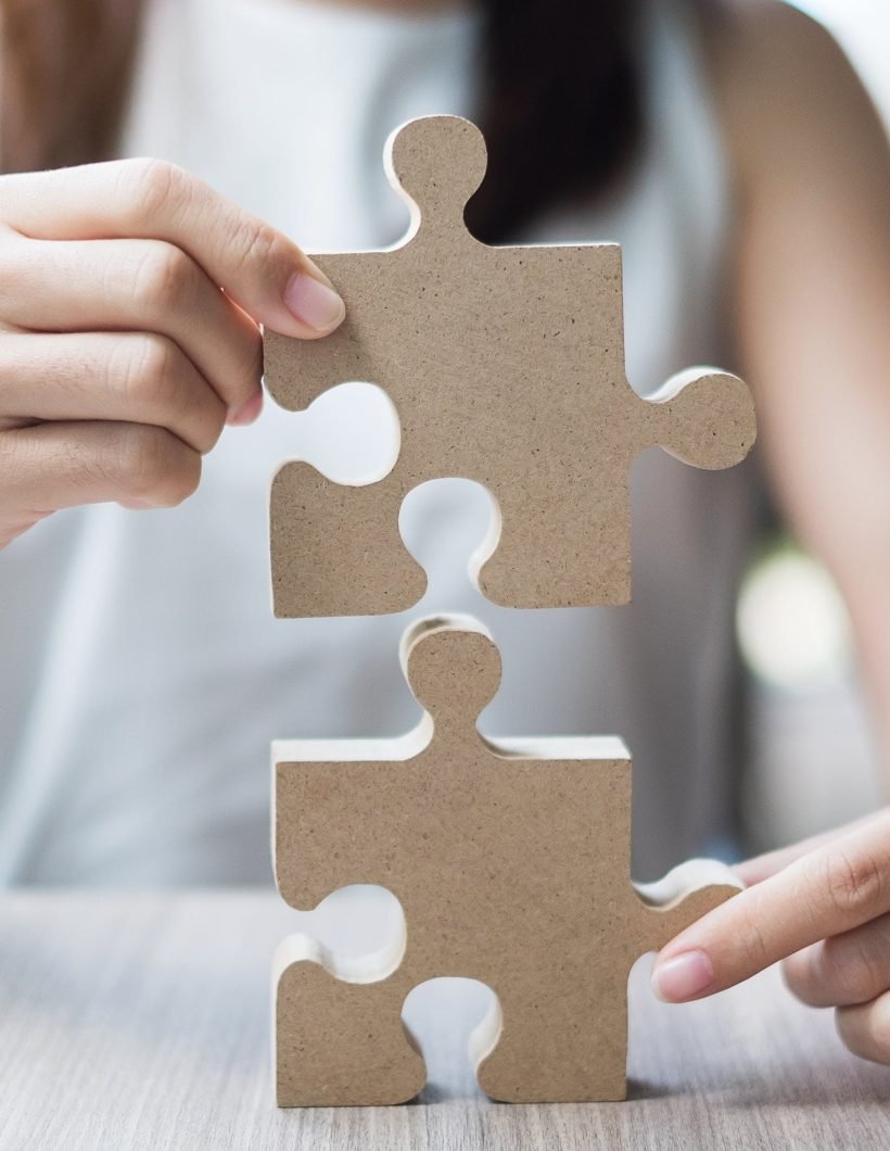 Woman hands connecting couple puzzle over table, businessman holding wood jigsaw inside office